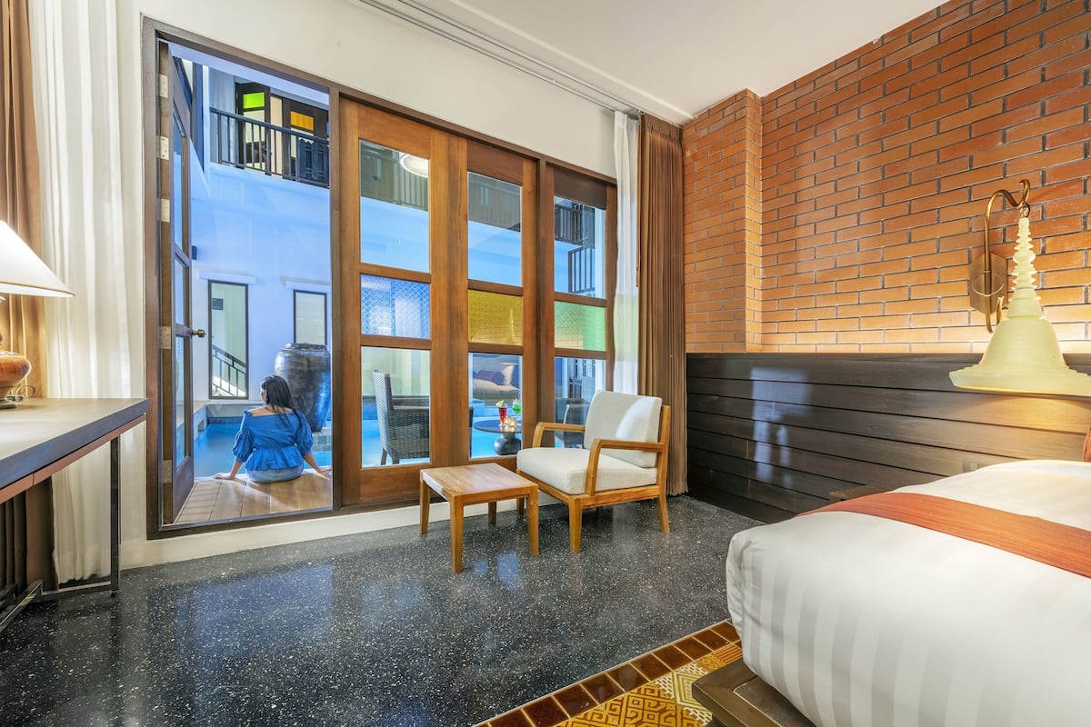 Quarto de hotel com uma grande porta de vidro deslizante que dá para uma varanda, uma cama com manta listrada, uma cadeira de madeira e uma pequena mesa.