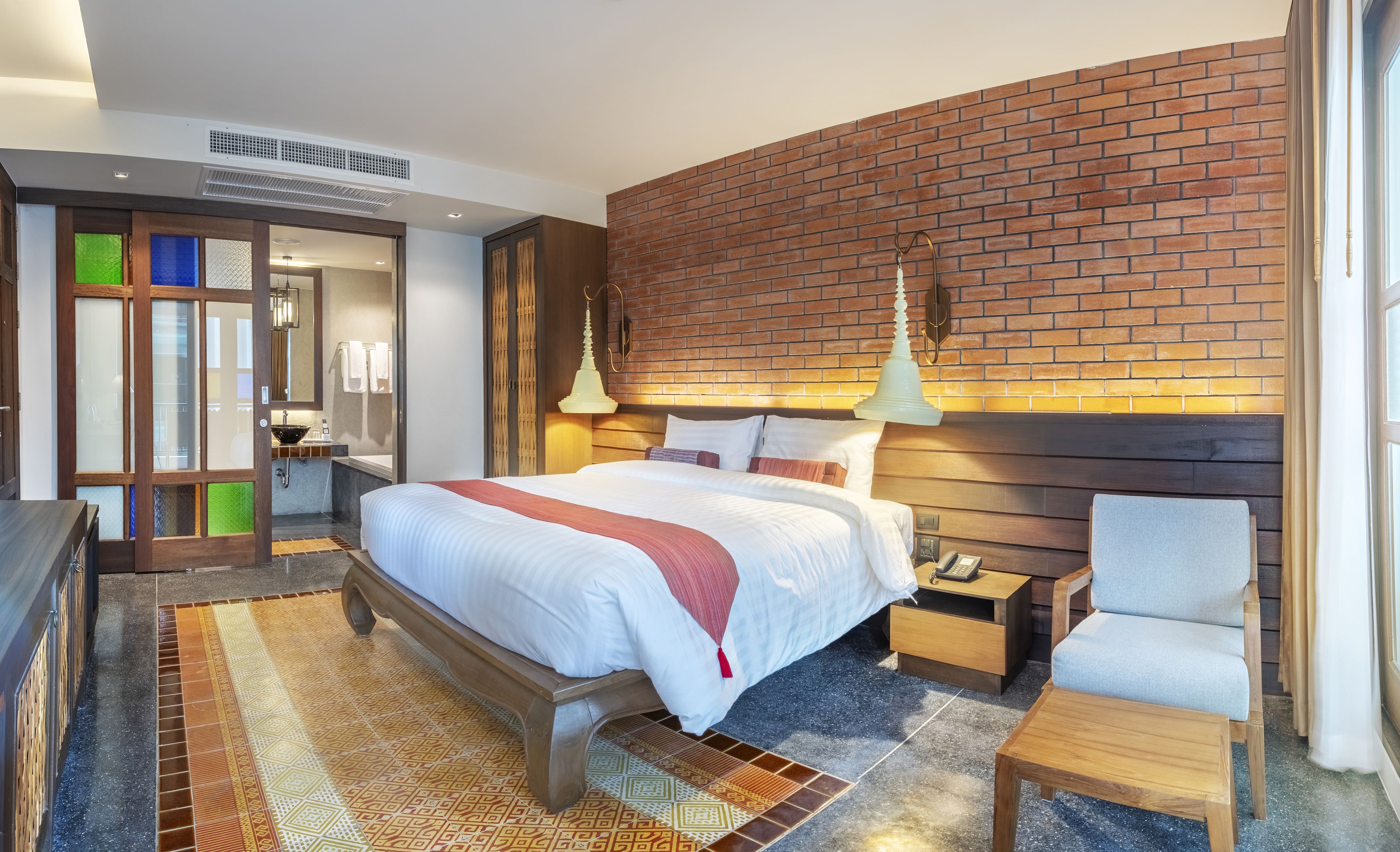 Una habitación de hotel moderna con una cama grande, una mesita de noche de madera, una silla, un baño con espejo y lavabo, y una pared de ladrillo decorativa.