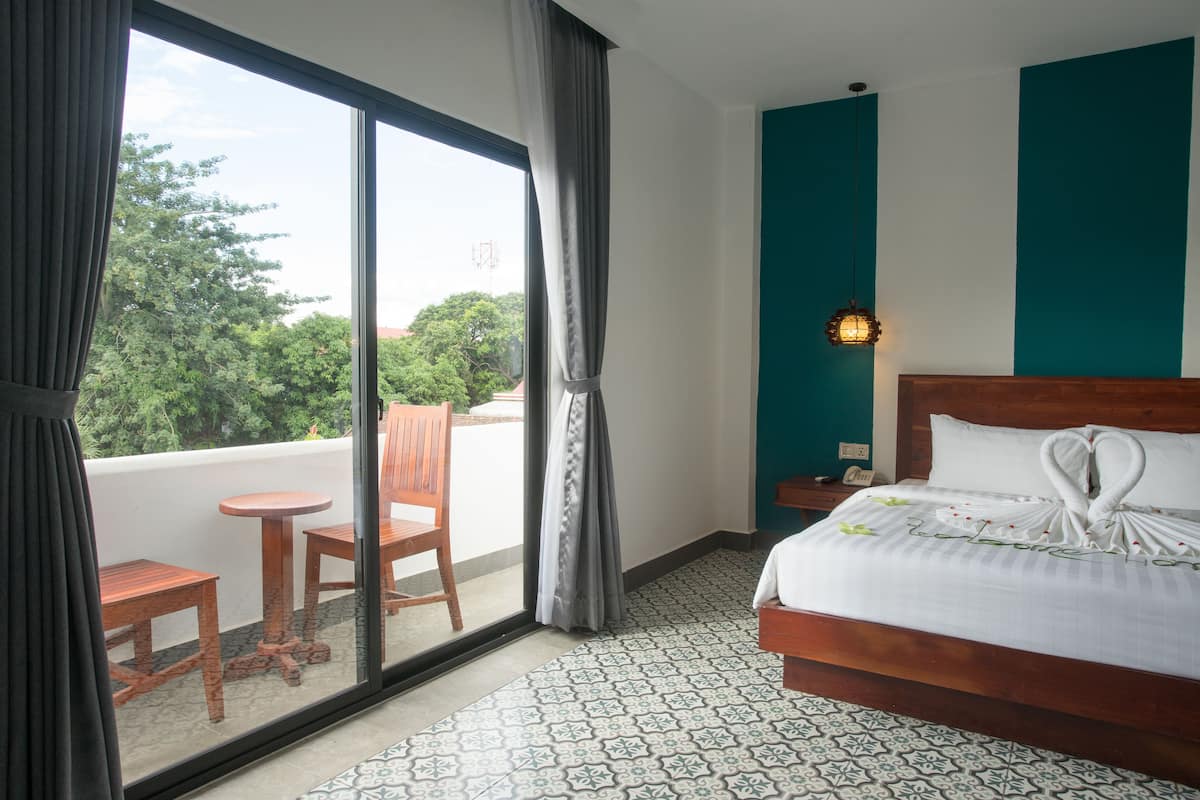 Een hotelkamer met een bed, een nachtkastje, een raam met gordijnen, een klein tafeltje en stoel, en een vloer met patroon.