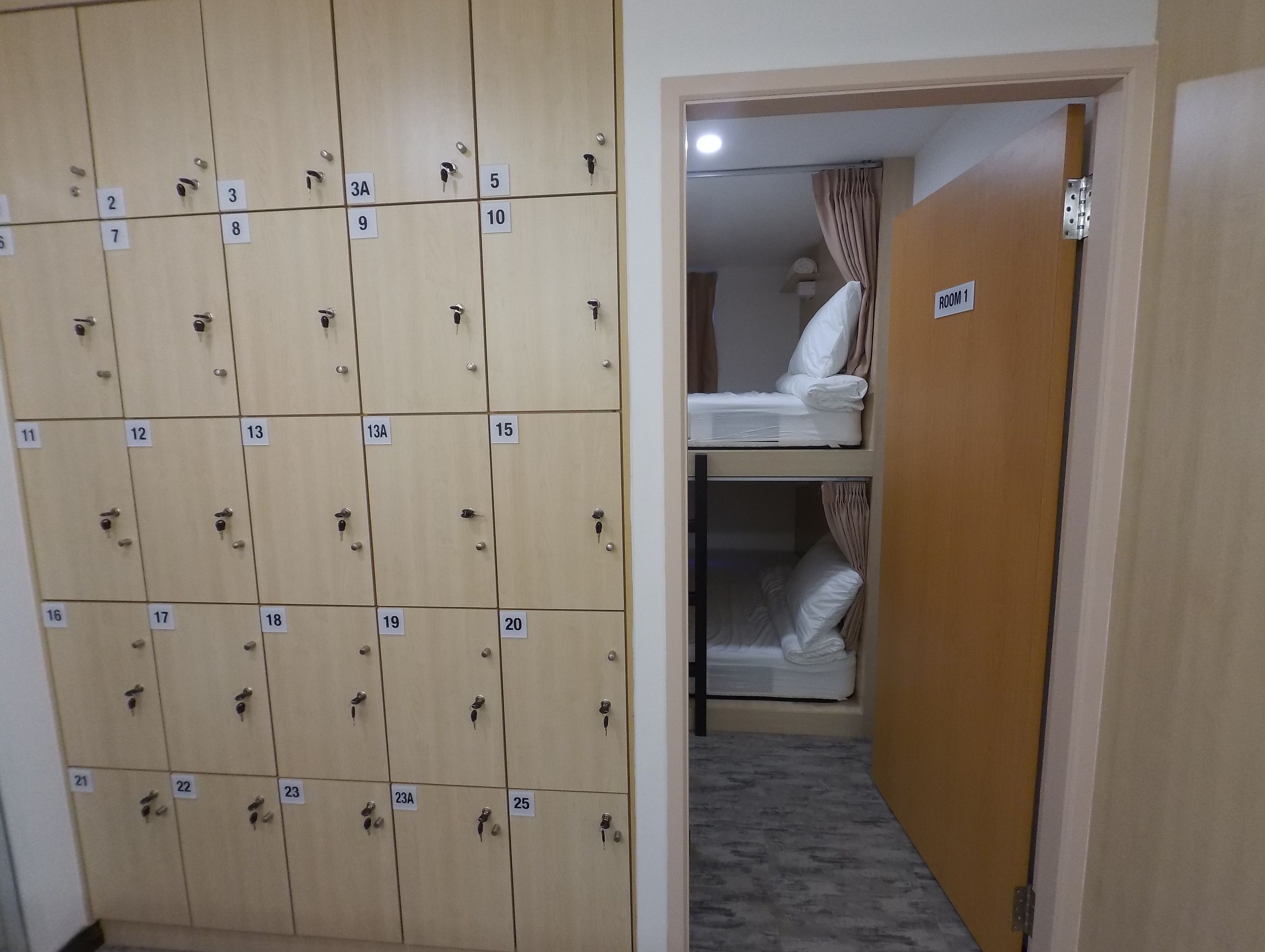 A hotel room with a large wooden wardrobe containing multiple compartments, each marked with a number. There is a door leading to a room with two beds, one on the upper and one on the lower bunk.