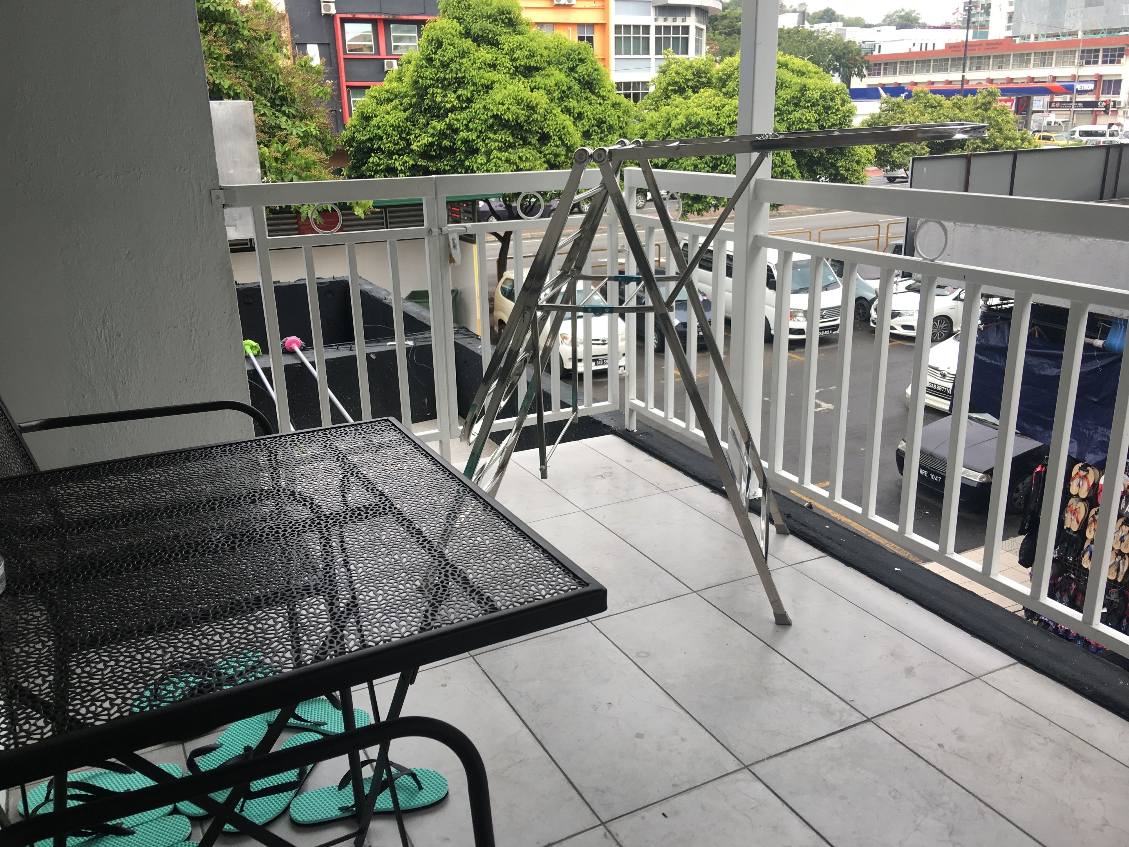 A balcony with a metal table and chairs, a view of parked cars, and a building with greenery in the background.