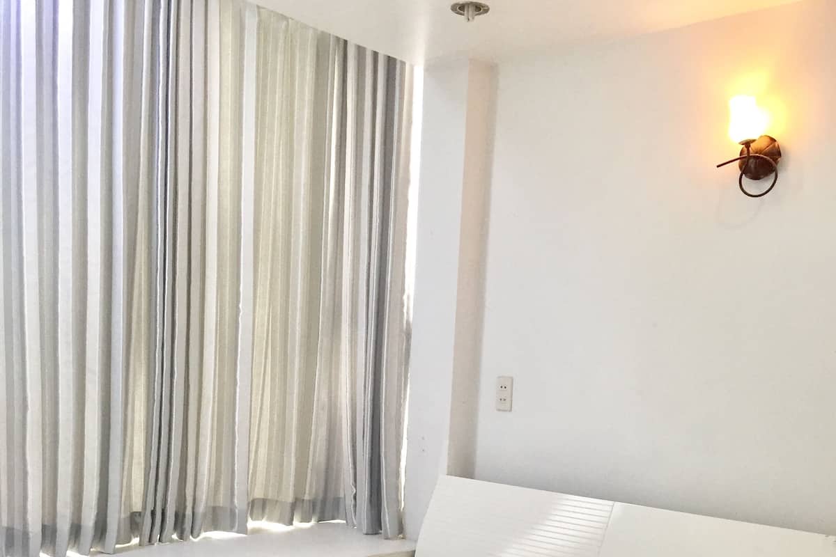 A neatly made bed with white linens and pillows, a window with sheer curtains, and a wall-mounted light fixture.