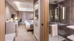 Une chambre d’hôtel moderne équipée d’un bureau, de deux lits, d’un espace douche et d’une salle de bain fermée par des parois de verre.