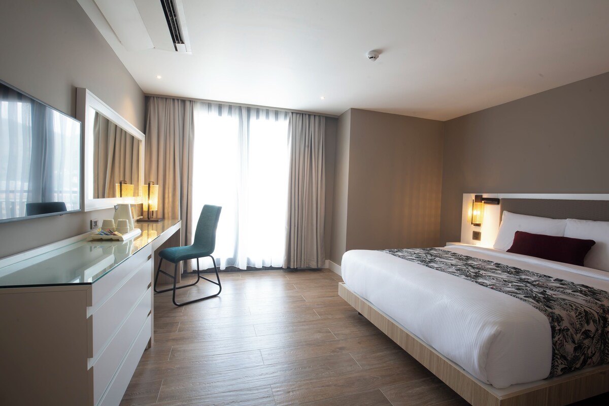 A modern hotel room with a large bed, a desk with a chair, a TV, and a window with curtains.