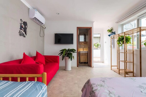 Una habitación de hotel moderna con un sofá rojo, una cama con colcha estampada y un televisor montado en la pared, además de una estantería de madera para plantas.
