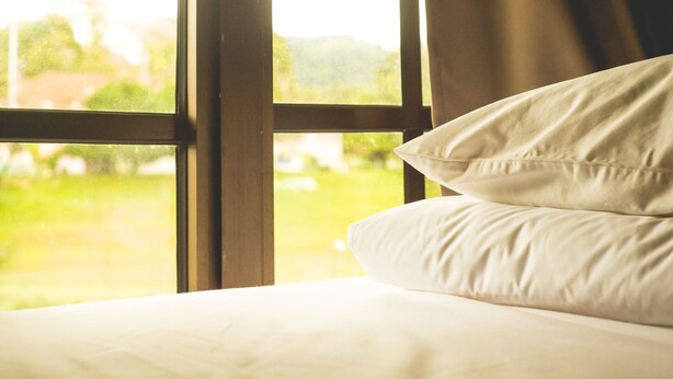 A bed with white bedding and pillows, positioned next to a window with a view of greenery outside.