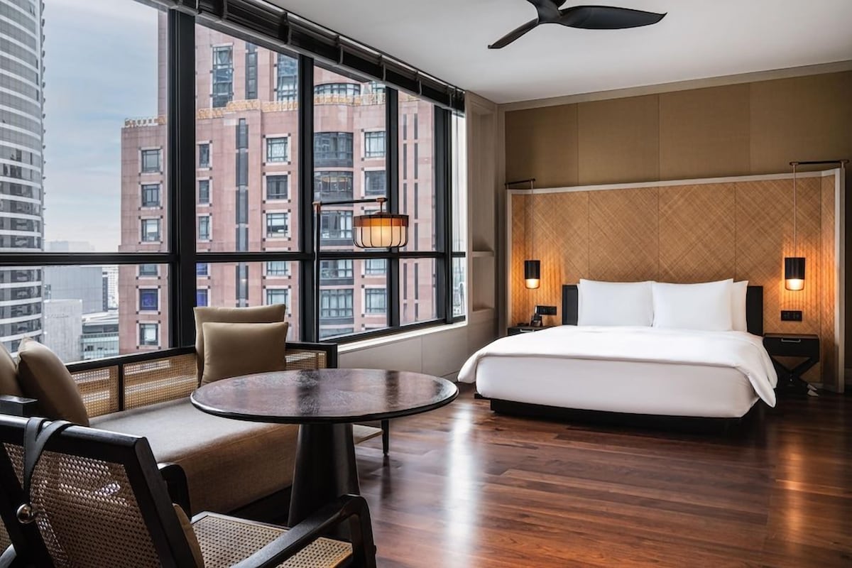 A hotel room with a bed, a round table, chairs, a large window with a city view, and a ceiling fan.