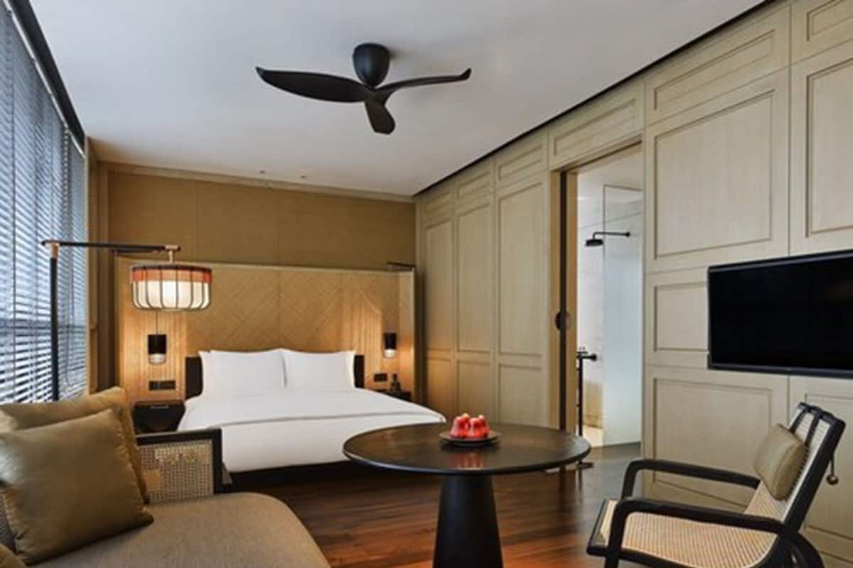 Une chambre d’hôtel avec un lit, une table ronde sur laquelle se trouve un objet rouge, un ventilateur de plafond, une télévision et une fenêtre avec des stores.