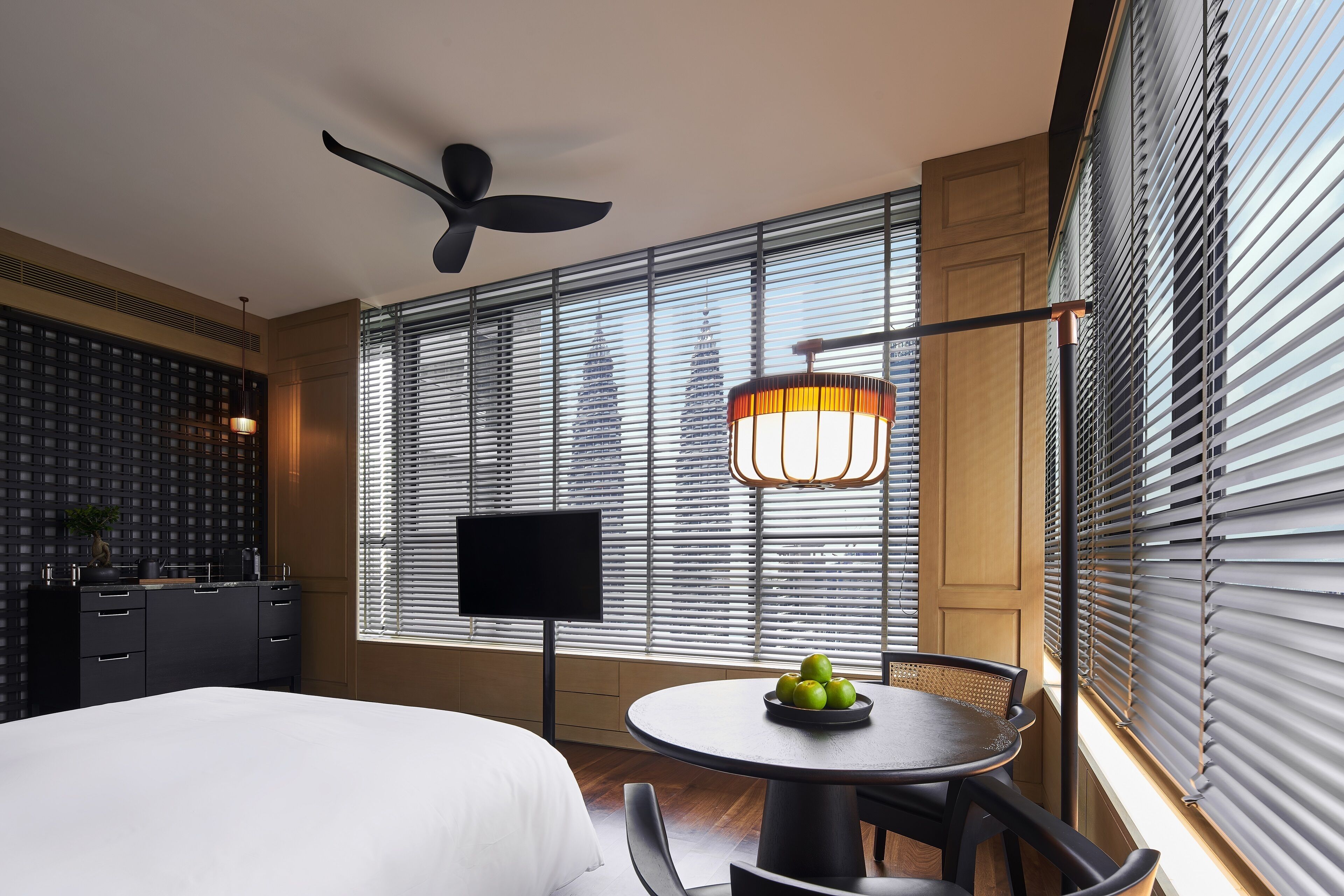 A modern hotel room with a bed, a round dining table with chairs, a ceiling fan, a TV, and large windows with blinds.