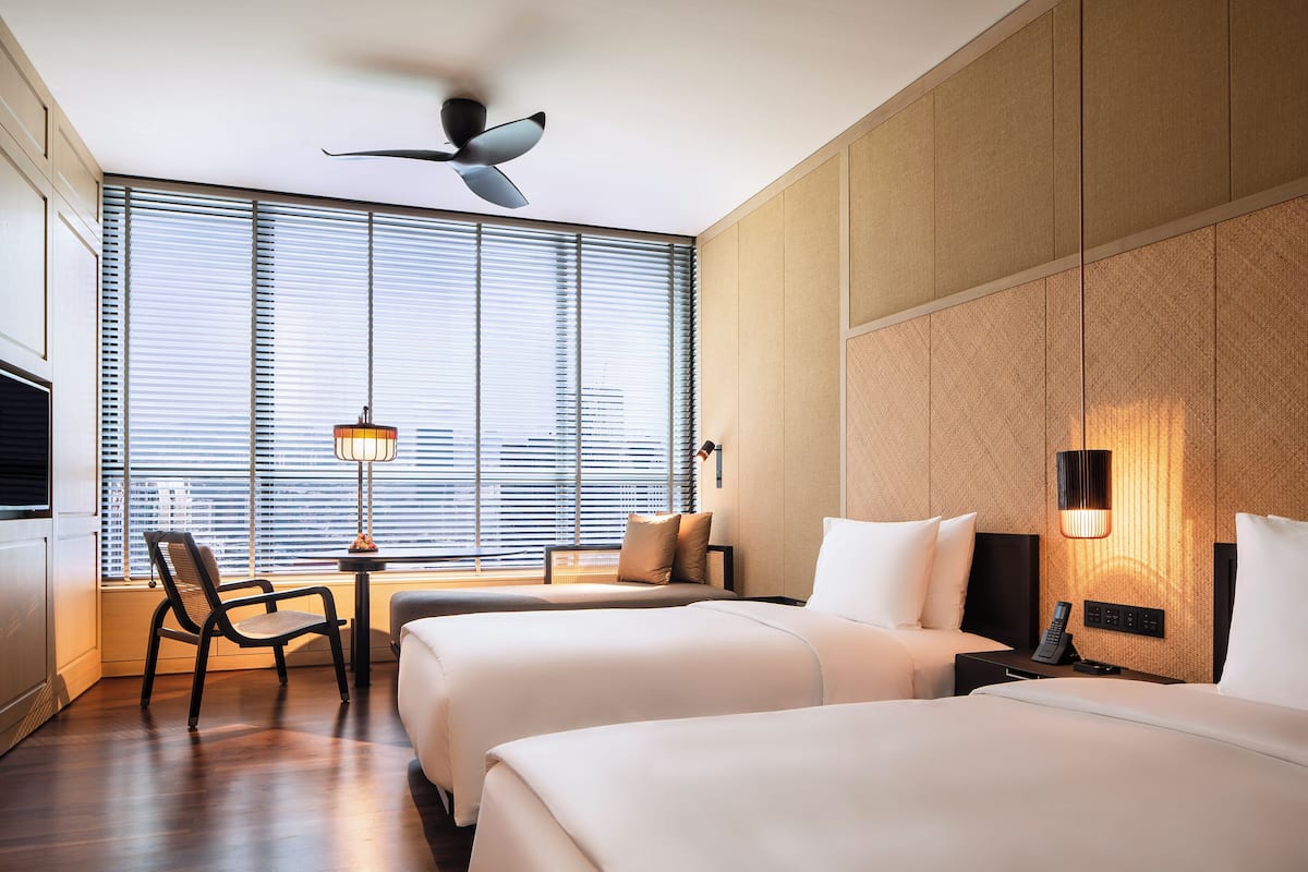 Une chambre d’hôtel avec deux lits, un ventilateur de plafond, une fenêtre avec des stores, un bureau avec une lampe et une chaise.
