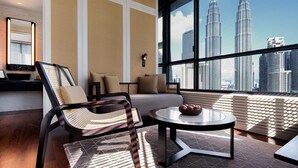 Une chambre d’hôtel moderne dotée d’une grande fenêtre offrant une vue sur les tours Petronas. La chambre comprend un canapé, une table basse ronde avec des fruits et un fauteuil en osier.