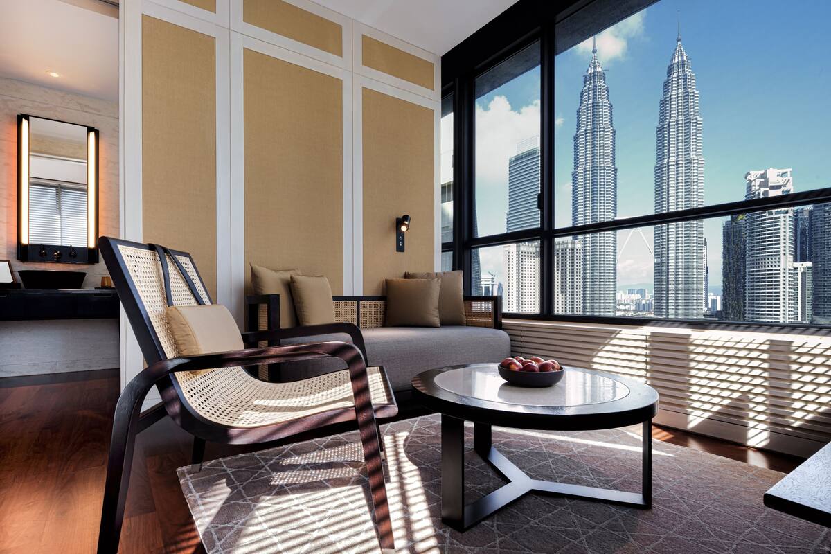 Une chambre d’hôtel moderne dotée d’une grande fenêtre offrant une vue sur les tours Petronas. La chambre comprend un canapé, une table basse ronde avec des fruits et un fauteuil en osier.