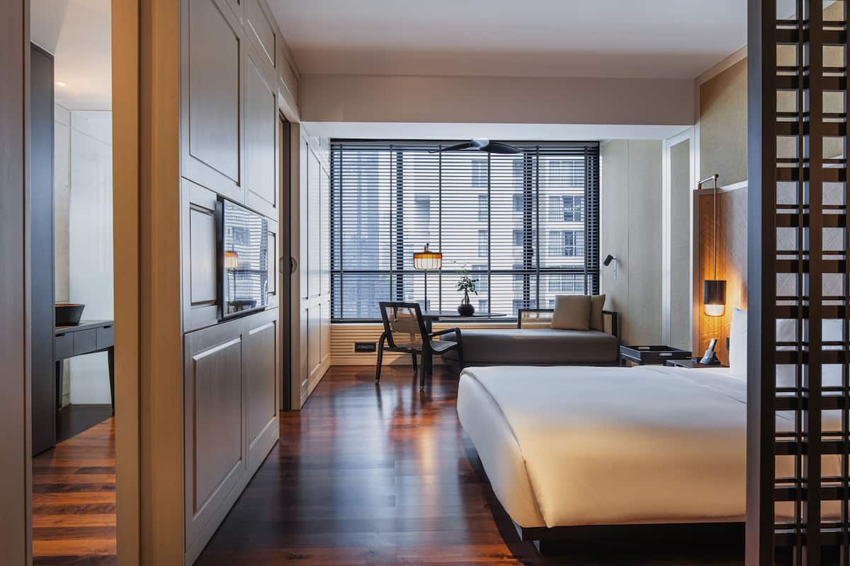Une chambre d’hôtel moderne dotée d’un grand lit, d’un bureau avec une chaise, d’une fenêtre avec des stores et offrant une vue sur la ville.