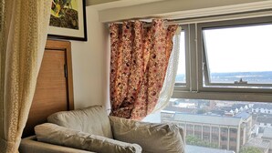 A room with a sofa, a small ottoman, a window with a view of buildings, and a curtain with a floral pattern. - Emma's home (Suzhou)
