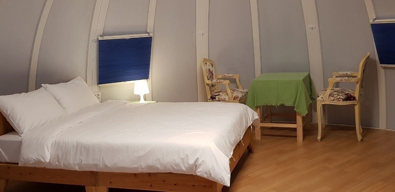 A bedroom with a wooden bed, two chairs, a small table with a green tablecloth, and a window with blue blinds.