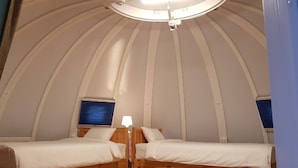 Una habitación abovedada con dos camas, una lámpara en el techo y dos ventanas con persianas.