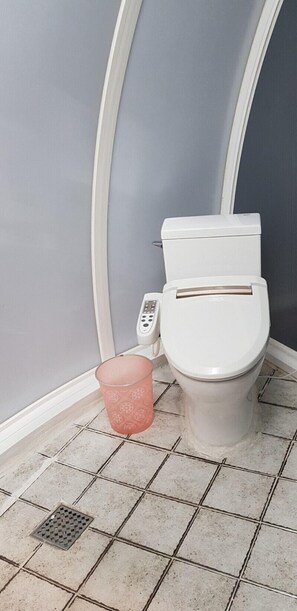 A bathroom with a toilet, a pink trash bin, and a floor drain. - Jeju Igloo Pension (Jeju City)