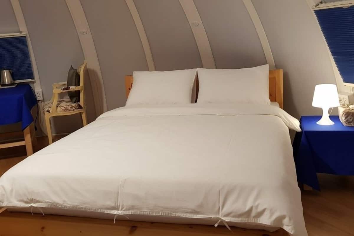 Een slaapkamer met een bed, nachtkastje met een lamp, een klein tafeltje met waterkoker en een stoel.