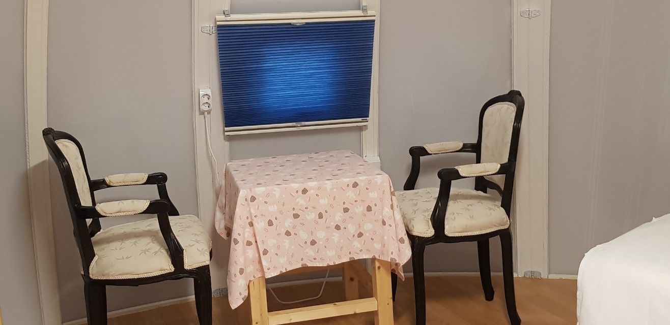 Une petite pièce avec une table recouverte d’une nappe à motifs floraux, deux chaises, une fenêtre avec des stores et un lit.