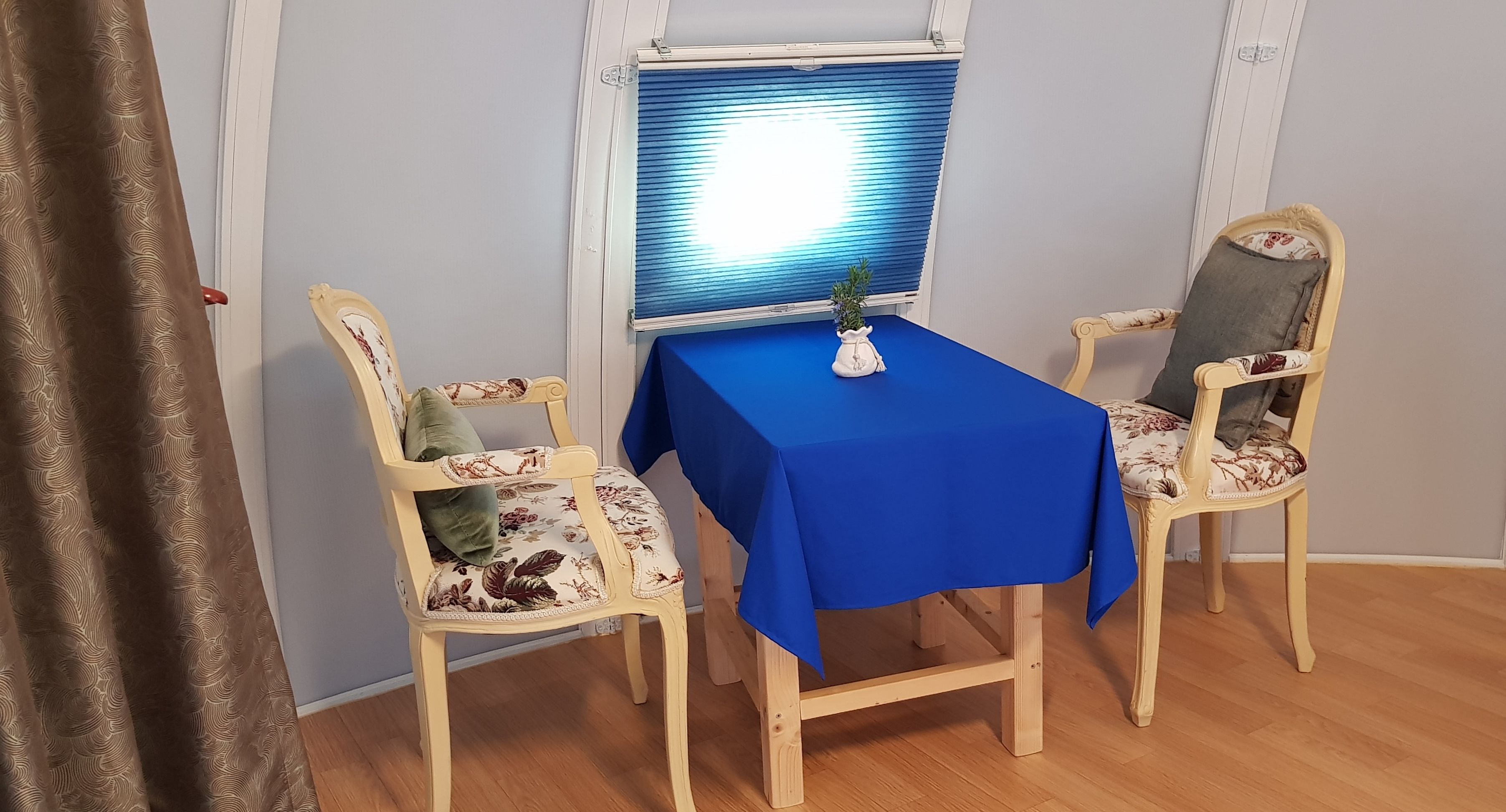 Un pequeño comedor con una mesa cubierta por un mantel azul, dos sillas con estampado floral y una ventana con persianas.