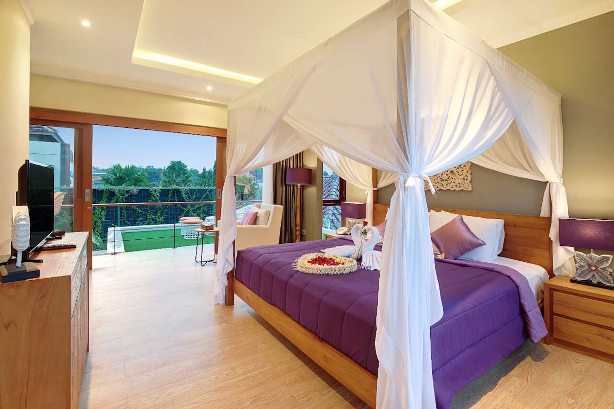 A bedroom with a large bed, a TV, and a balcony with a view of greenery.
