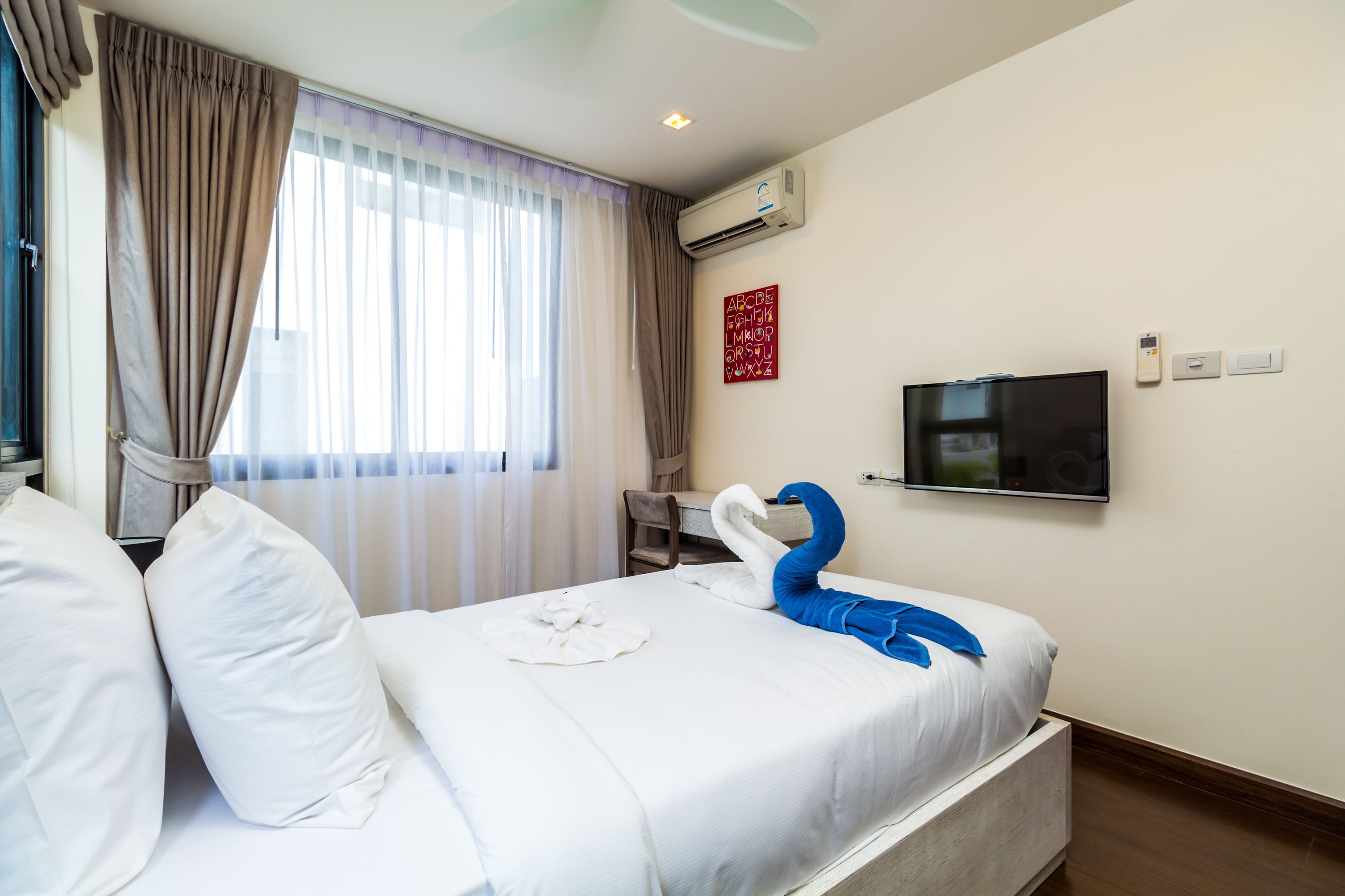 A hotel room with a bed, a flat-screen TV, an air conditioning unit, and a window with curtains.