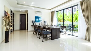 A modern dining area with a long table, chairs, and a view of greenery through large windows.