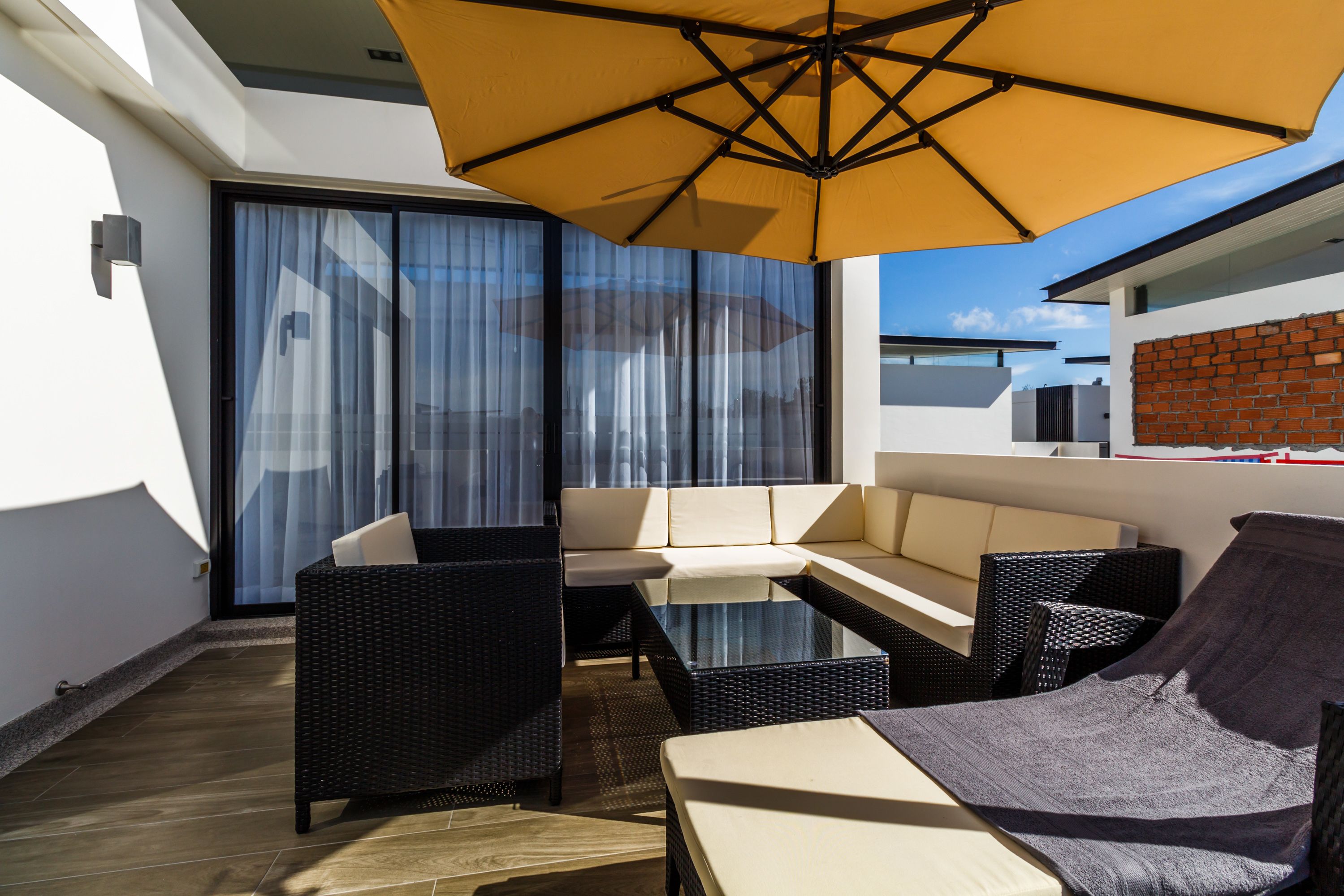 A modern outdoor seating area with wicker furniture, a glass table, and a large yellow umbrella.