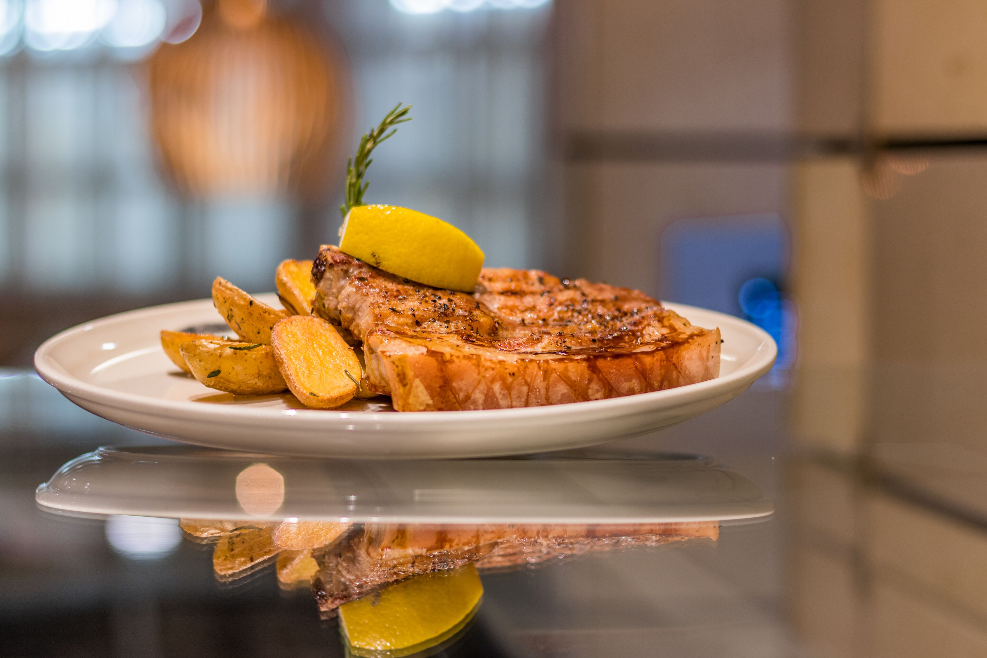 Un piatto con bistecca alla griglia, patate arrosto e spicchio di limone su una superficie riflettente.