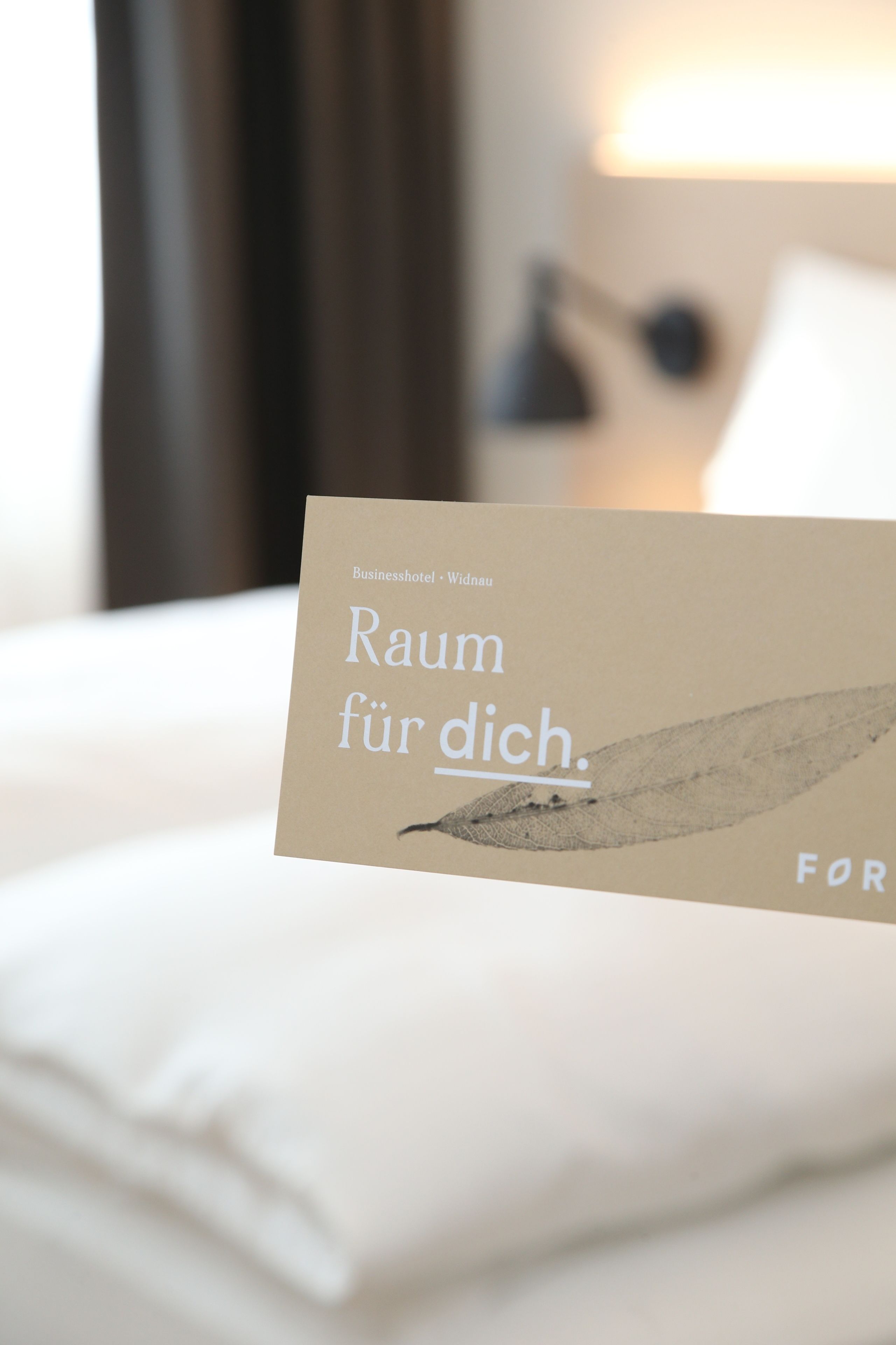 A hotel room with a neatly made bed, a lamp, and a card on the bed.