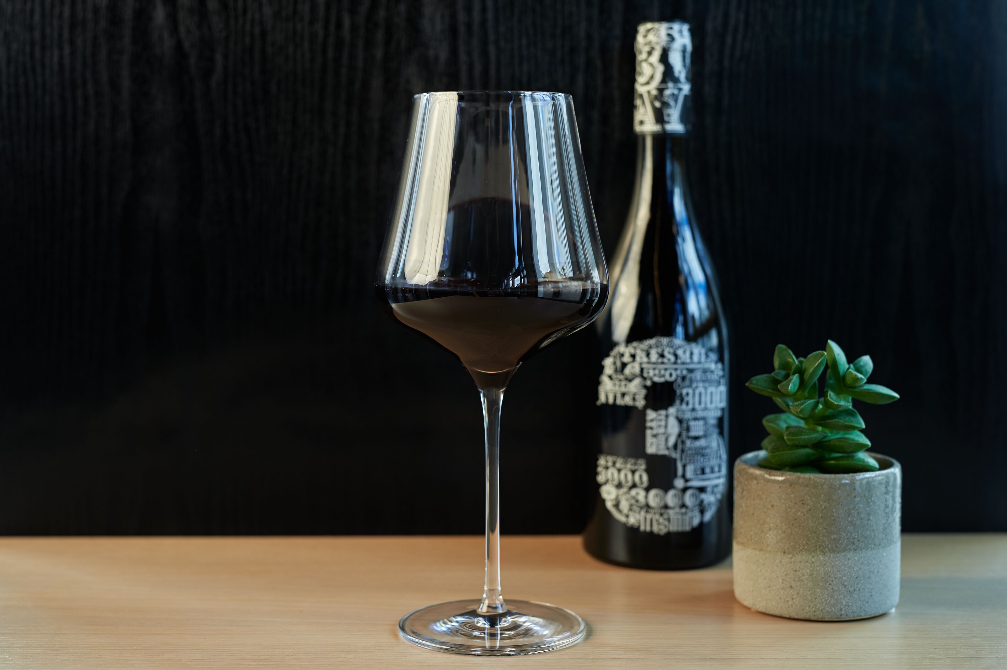 Ein Weinglas mit Rotwein, eine Weinflasche und ein kleiner Topfpflanze auf einer Holztischfläche.