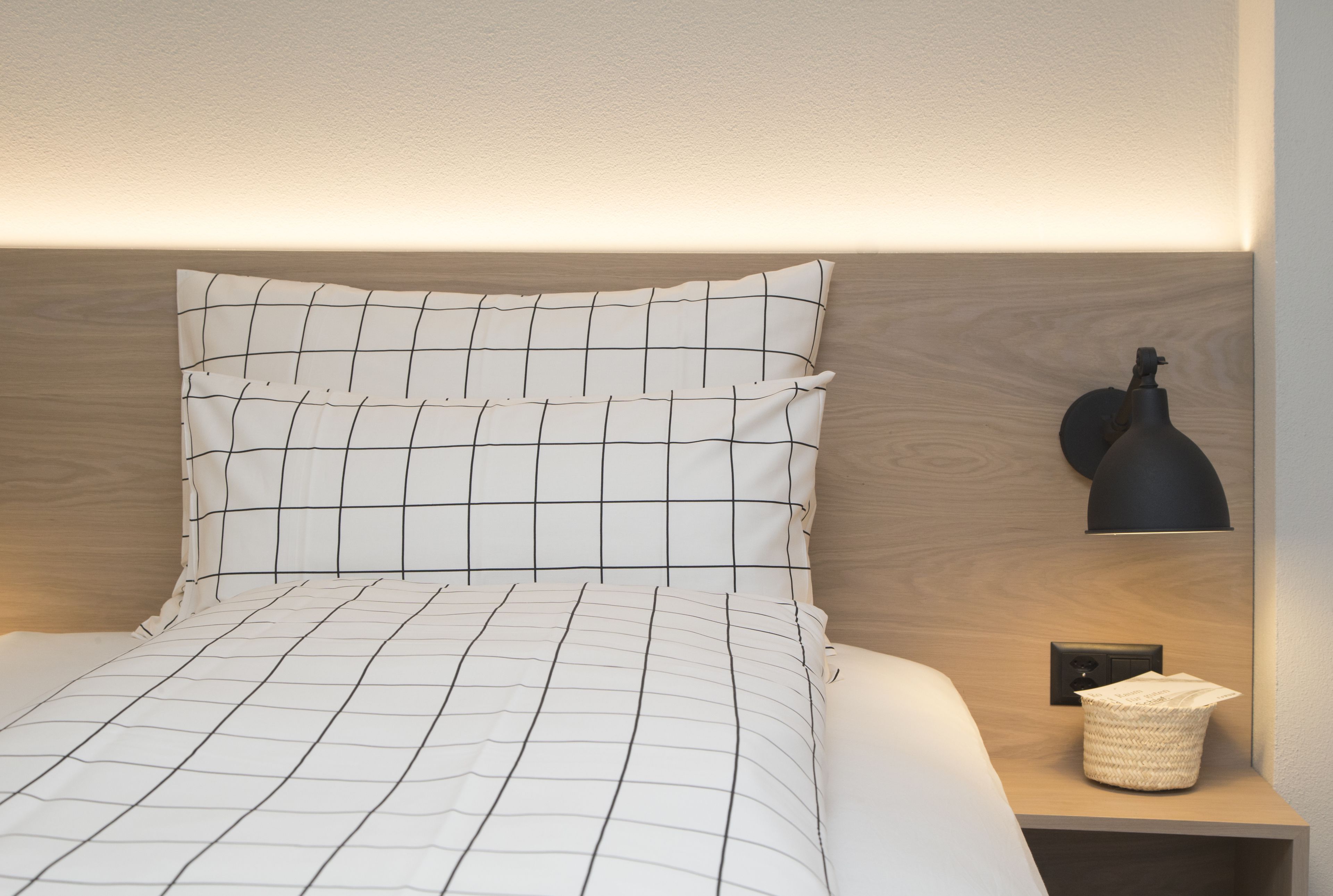 Una cama bien tendida con una colcha de diseño blanco y negro tipo cuadrícula, una lámpara negra sobre una mesita de noche y una cesta tejida.