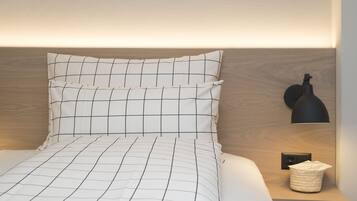 Una cama bien tendida con una colcha de diseño blanco y negro tipo cuadrícula, una lámpara negra sobre una mesita de noche y una cesta tejida.