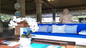 A blue sofa with patterned cushions, a wooden cabinet, and a Buddha statue in a covered area with a thatched roof. - The White Heron (Ubud)