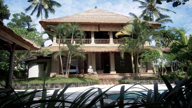 A two-story building with a thatched roof, a balcony, and a statue in front. - The White Heron (Ubud)