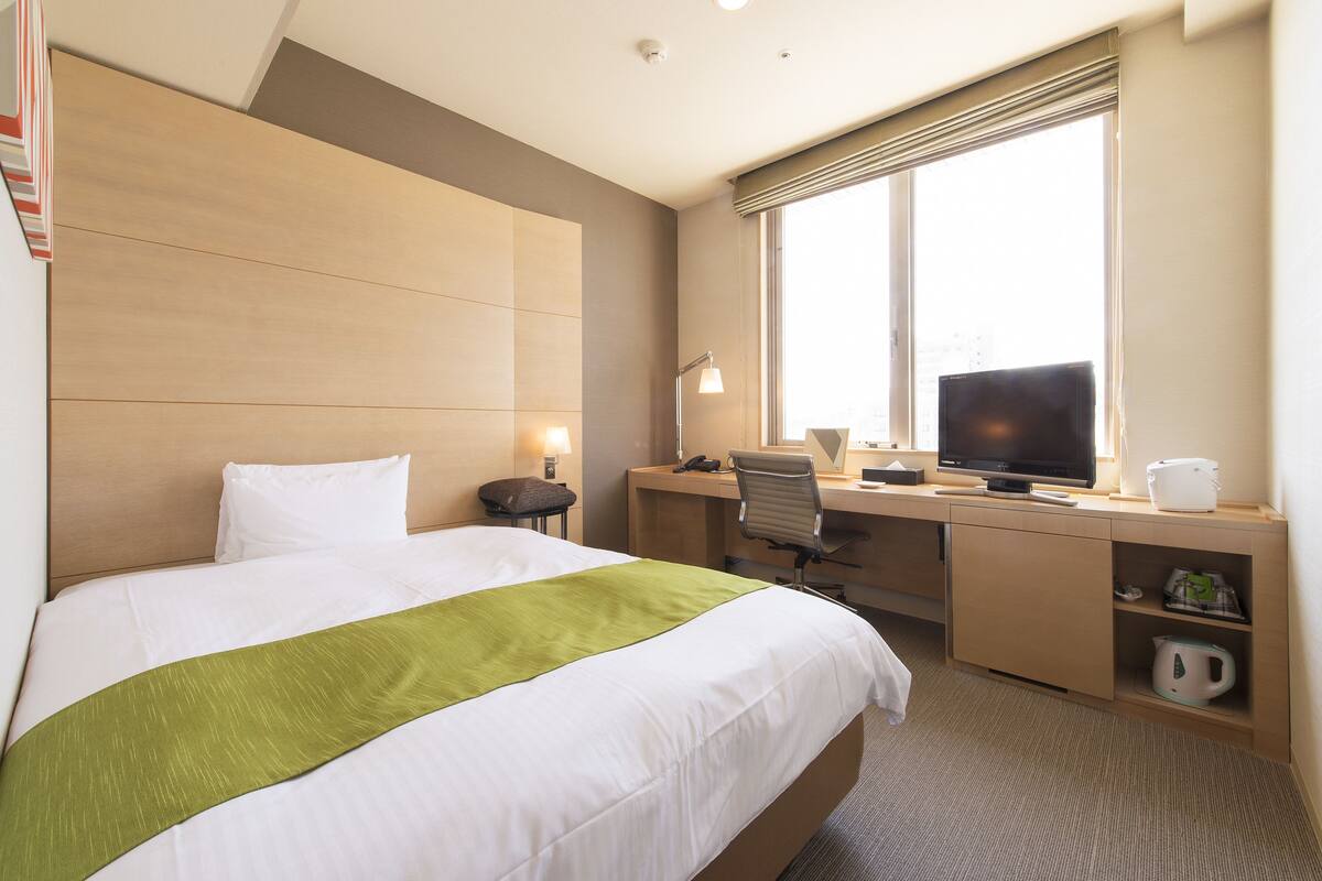 Een hotelkamer met een bed, een bureau met een computer, een televisietoestel en een raam met zonwering.