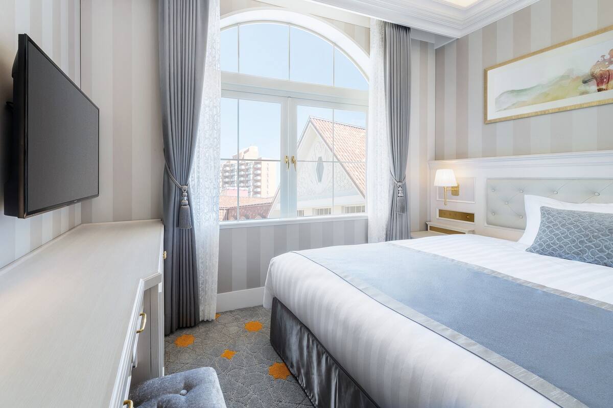 A hotel room with a large bed, a flat-screen TV mounted on the wall, a window with curtains, and a painting on the wall.
