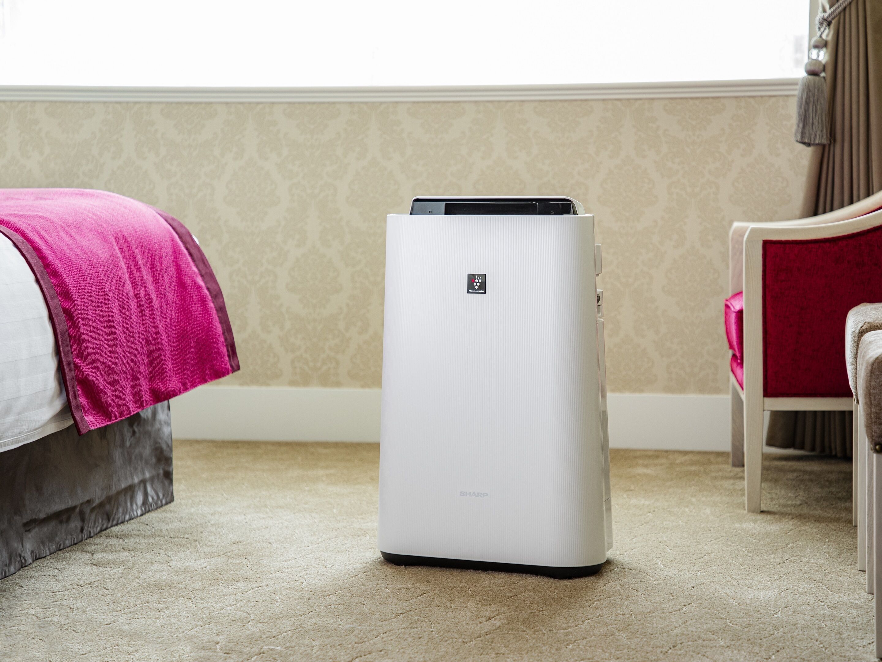 A white portable air purifier in a hotel room with beige patterned wallpaper, a bed with a pink towel, and a red chair.