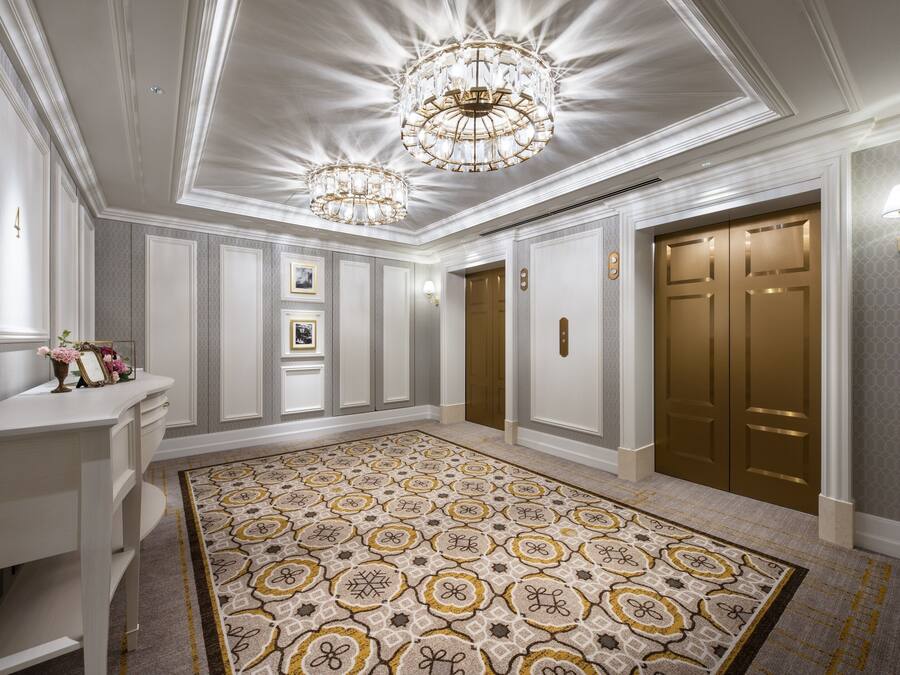 A spacious hallway with a patterned carpet, elegant chandeliers, and a set of double doors.