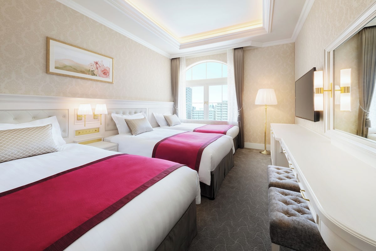 A hotel room with three beds, a large window, a TV, and a bench.