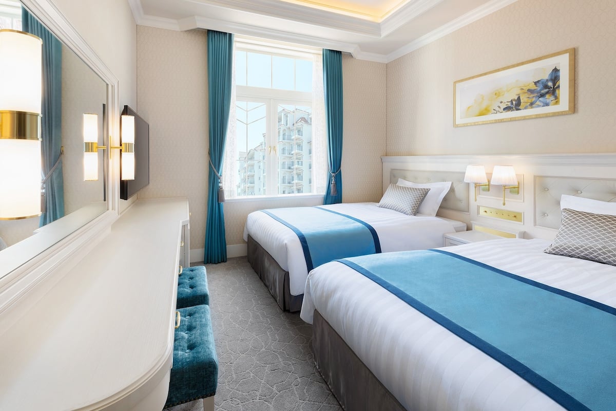A hotel room with two beds, a large window with curtains, a mirror, and a painting on the wall.