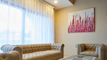 A living room with a beige sofa, a matching armchair, and a glass coffee table. A large abstract painting is on the wall.
