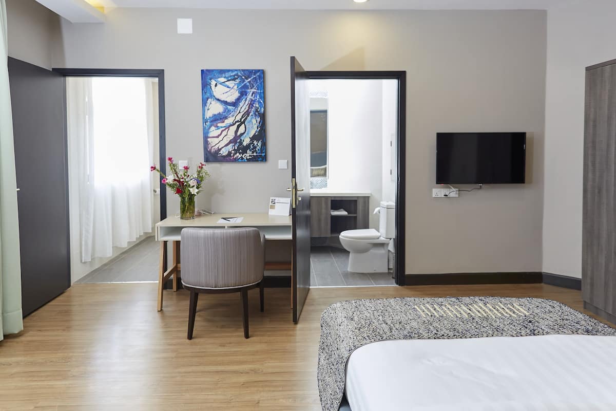 Una habitación de hotel moderna con cama, escritorio con silla, un cuadro, televisor y baño visible a través de una puerta abierta.