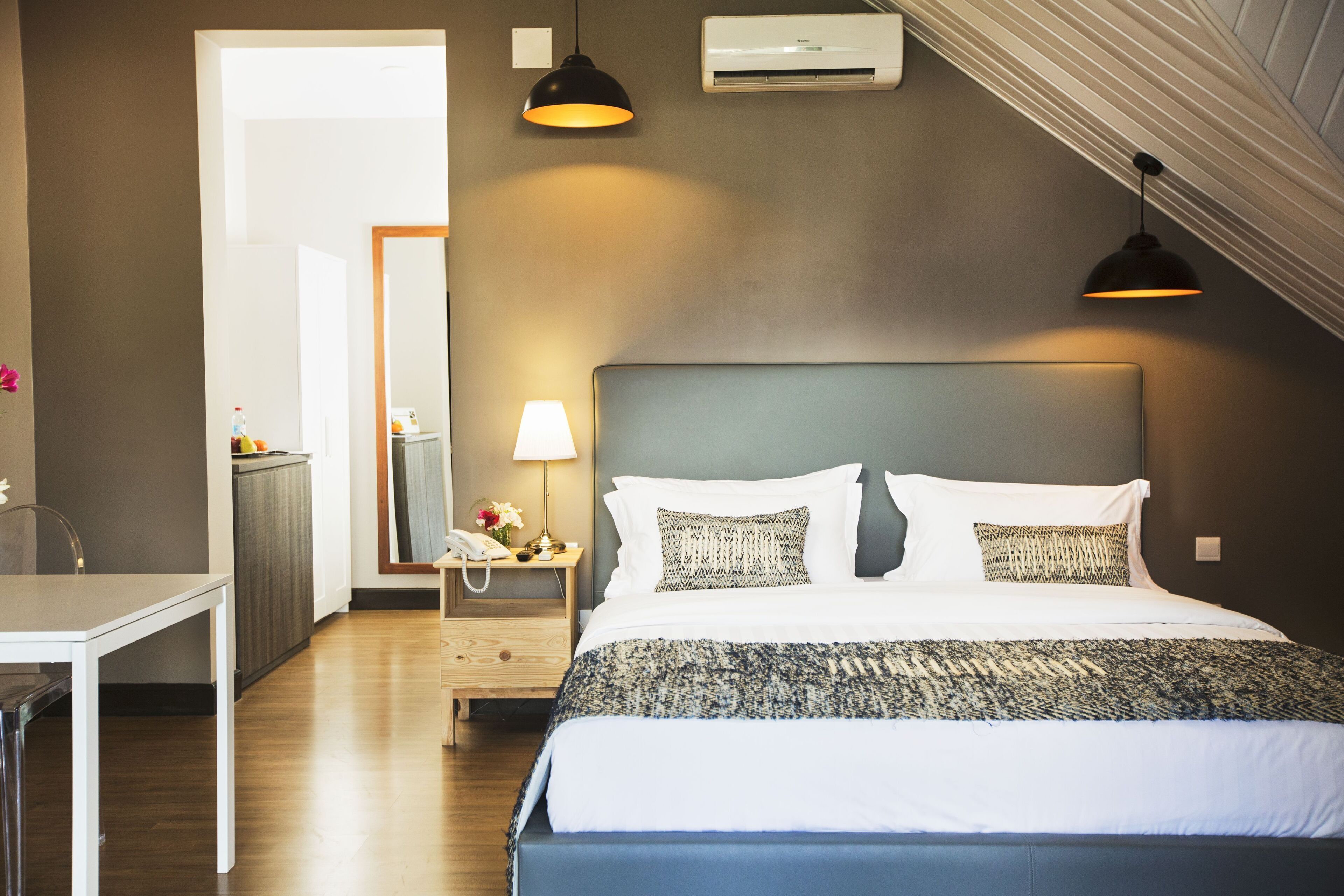 A hotel room with a bed, bedside table, lamp, and a view of a hallway.