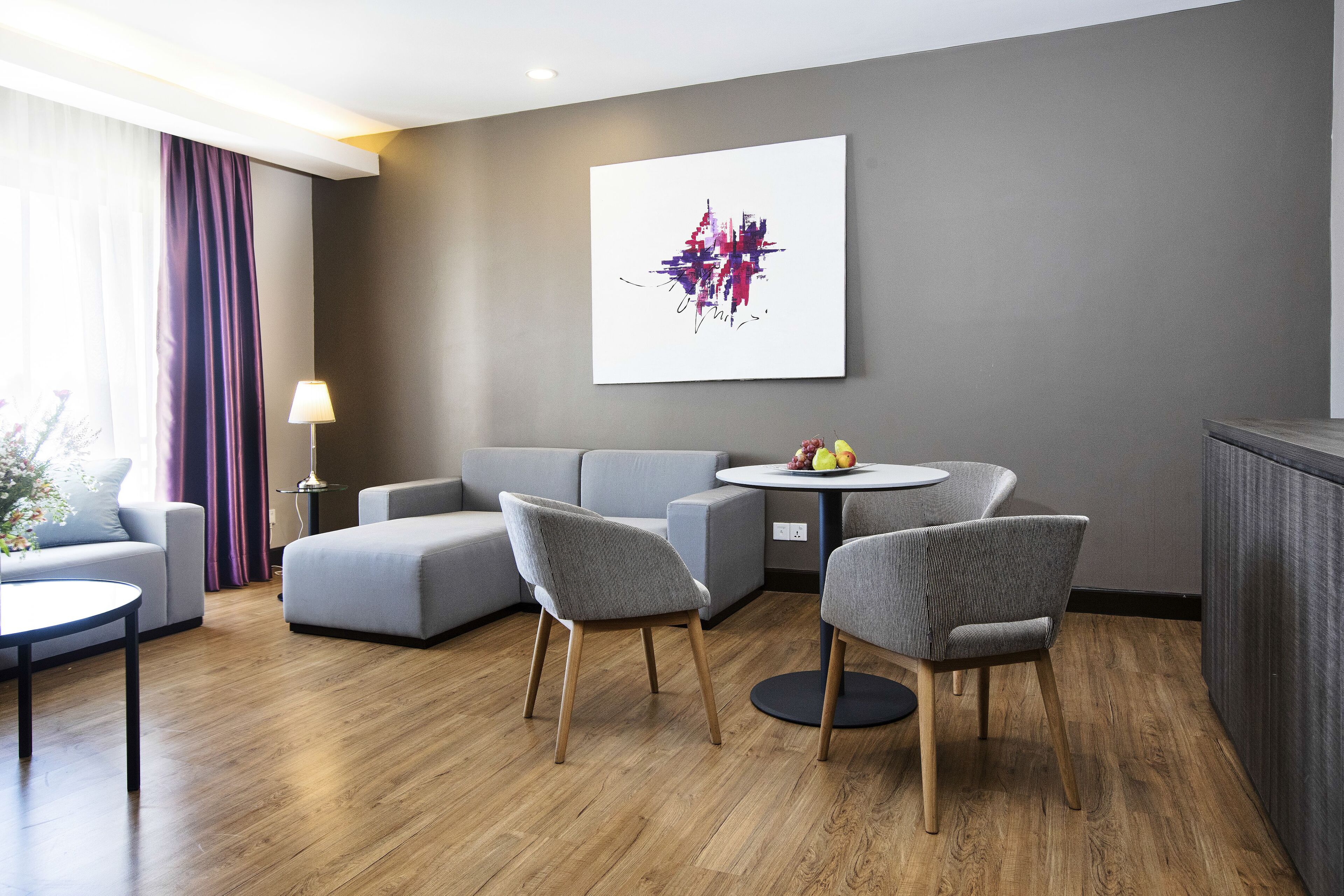 Una habitación de hotel moderna con sofás grises, una mesa de comedor redonda y un cuadro abstracto contemporáneo en la pared.