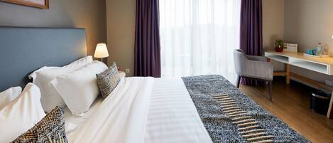 A hotel room with a bed, desk, chair, and a painting on the wall.
