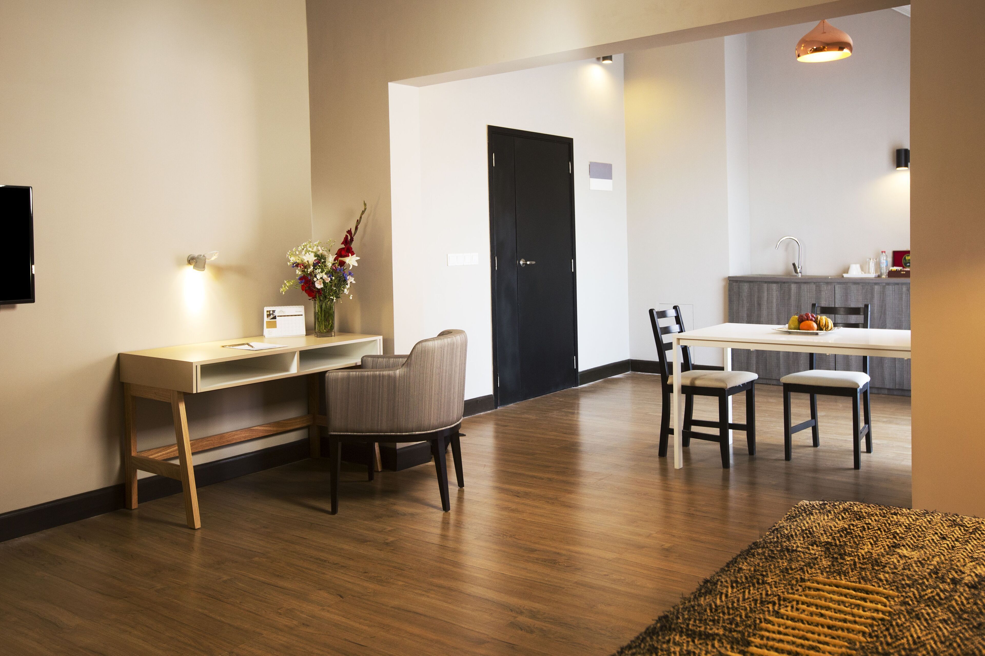 A modern living room with a wooden floor, a desk with a chair, a dining table with chairs, and a kitchen area in the background.