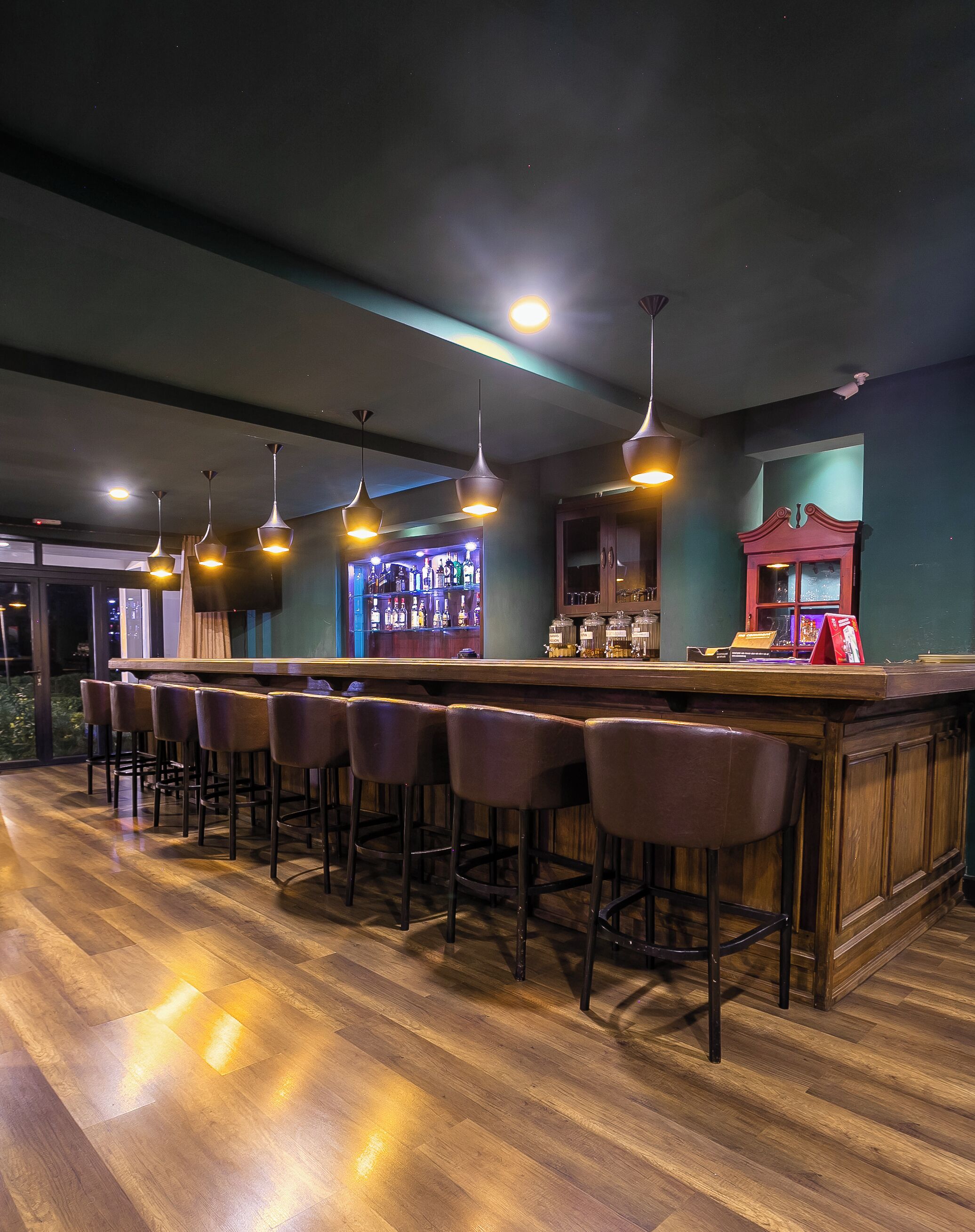 A bar area with a long counter, multiple stools, hanging pendant lights, and a bar stocked with bottles.