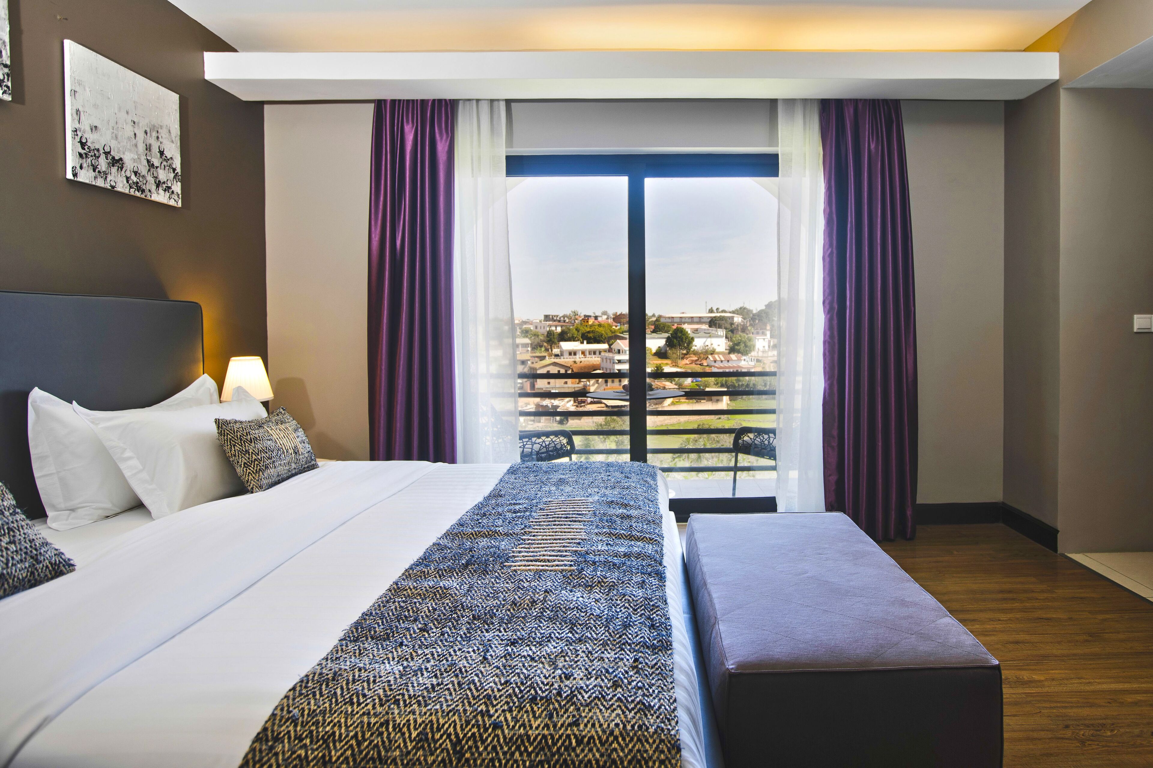 A modern hotel room with a large bed, a bench, a view of the city through a balcony, and a cityscape visible through the window.