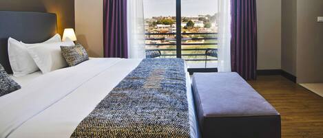 A modern hotel room with a large bed, a bench, a view of the city through a balcony, and a cityscape visible through the window.